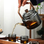 Glass teapot with wooden handle being poured into glass cups on a wooden tray.